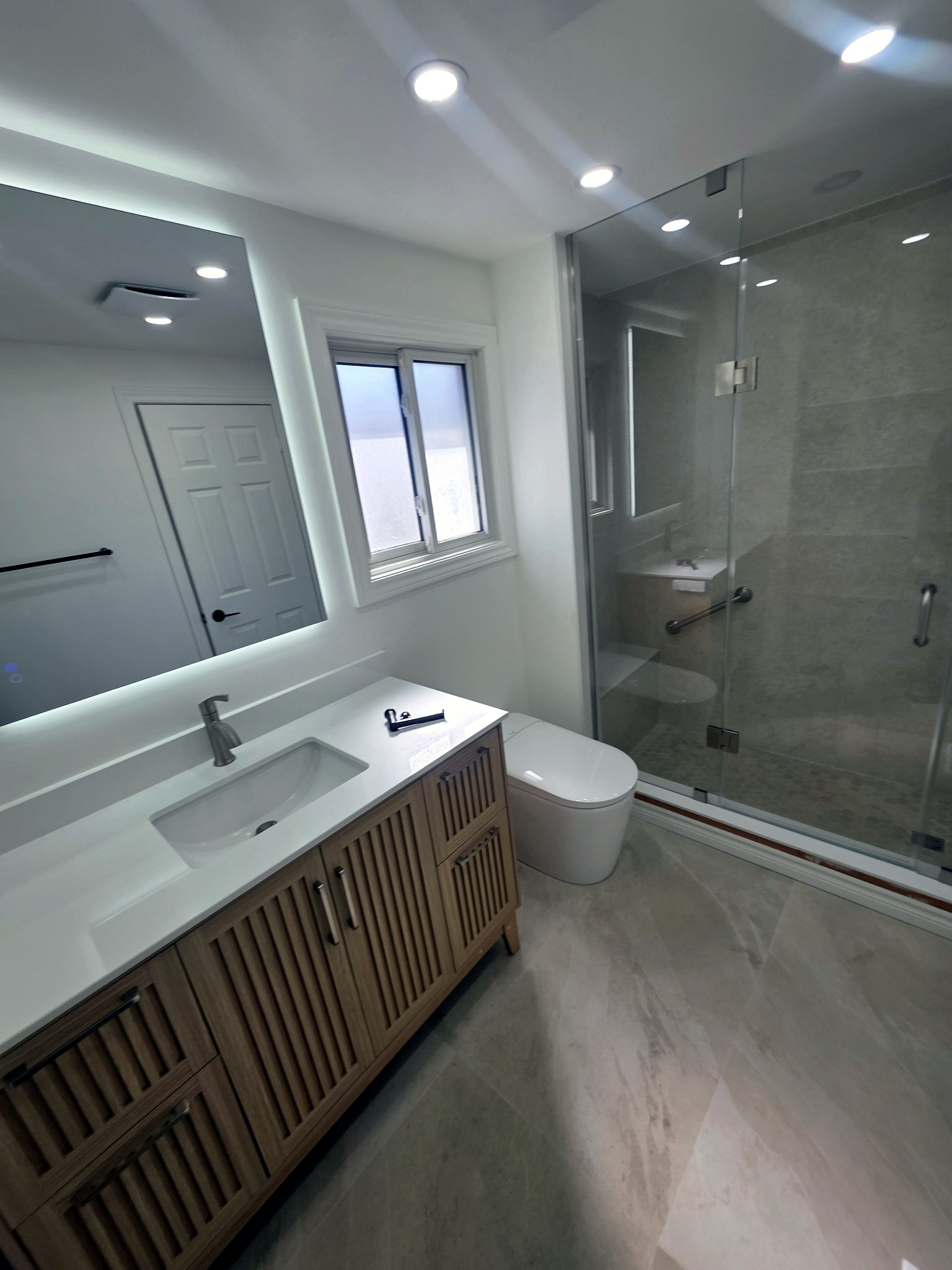 Walk-in glass shower with stone tile and bench beside wood vanity